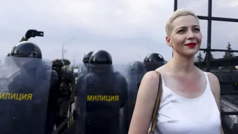 Getty Images Maria, wearing a white singlet and with short blonde hair, smiles as she stands in front of officers in riot gear with sheilds.