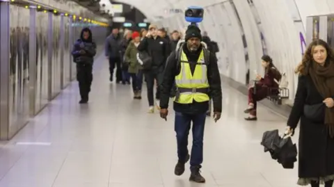 Man in yellow high vis jacket carrying a blue street view camera on his back walking down a Tube platform