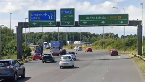 Several lanes of traffic approaching Switch Island with signs for the M57 Liverpool Airport, A59 Liverpool and Bootle docks A5036