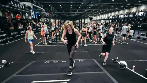 Participants at a Hyrox event. They're running and lifting and throwing weights inside a gym space. 
