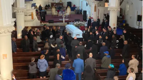 Mr Meade's white coffin is brought in to the church with mourners behind and standing in the pews.