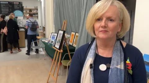Karen Worrall stands in front of an easel holding a colourful artwork in a black frame. Behind her are other people, one wearing black and another in black trousers and a blue striped shirt, queueing for a cafe. Karen is a white woman with short blonde hair and is wearing a blue jumper with a scarf in an abstract white and blue pattern, she also has a necklace of coloured circles. She is also wearing a red poppy brooch. 