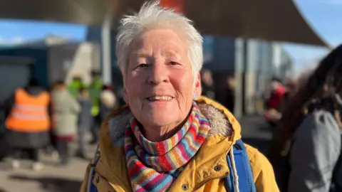 George Carden/BBC A woman in a yellow jacket and a colourful scarf, smiling at the camera.