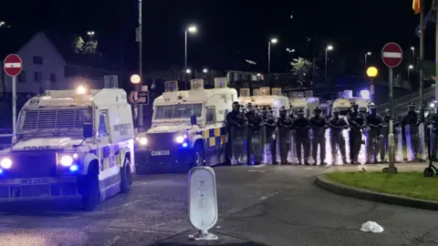 Sean Campbell Shows white police Land Rovers with more than a dozen riot police in black and wearing helmets and carrying transparent shields
