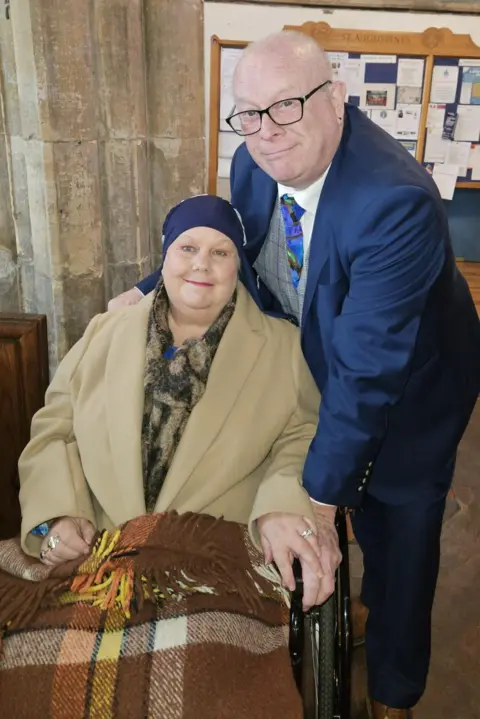 Family handout Donna and Sid pose together inside a church interior with stone pillars and stained‑glass windows. Donna is seated in a wheelchair, wearing a light-coloured coat and navy head covering, with a patterned blanket draped over her lap. Sid stands slightly behind the wheelchair, wearing a navy suit with a patterned tie and glasses, one hand is resting on the wheelchair handle. In the background, there is a wooden noticeboard displaying various papers and announcements.