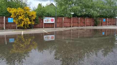 Emma/BBC Weather Watchers The puddle covers a large area of Tarmac next to a long wooden fence with trees behind it. A sign is reflected in the surface of the puddle.