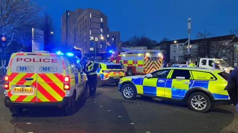 BBC Police and fire vehicles blocking the road close to the bus station. Three police vans and a fire engine in the foreground with flats in the background. It is twilight and the vans have their lights on