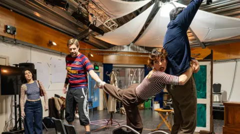 Michael Berkeley Four actors in a large rehearsal room with a rough scene set up including a door, a coat stand and four dining room chairs. Three of them are standing on the chairs with one reaching upwards and another clinging on to his waist with her leg being pulled by another man