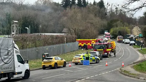 Police blocking the A46, as seen from Culver Hill. Three fire engines can be seen on the road. Some light smoke is visible in the background.
