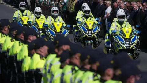 PA Media Seven Garda motorcycle police officers ride along the road in yellow and blue bikes and yellow high-viz jackets while around 15 other uniformed Gardai, also wearing yellow tops, stand to attention at the side of the road.
