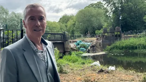 A smiling David King looking straight at the camera. He is wearing a grey suit and is standing on grass in front of brickwork which has collapsed into a river.