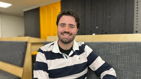A man with short, curly dark hair and a dark beard. He is staring directly at the camera and smiling, his teeth visible. He is wearing a top with chunky blue and white horizontal stripes. He is sitting indoors, on a bench made from wood with a grey, fabric, padded cover. Behind him, the wall is both yellow and grey.