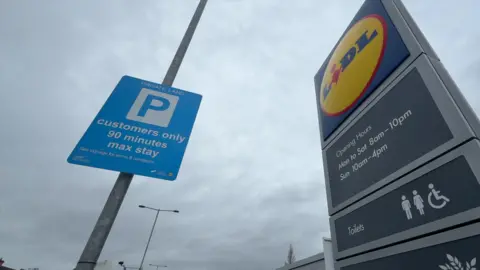 Two signs in a supermarket car park. A blue rectangular sign on the left of the image has been fixed to a lamp post and reads "Private land, customers only, 90 minutes max stay". Another sign on the right of the image features a blue, yellow and red Lidl logo and has details of the store opening hours. Both signs are set against a cloudy background.
