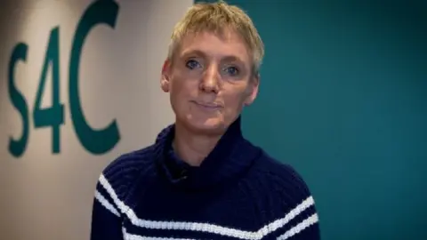 Sian Doyle wears a black jumper with two horizontal white stripes. S4C is written on the wall behind.