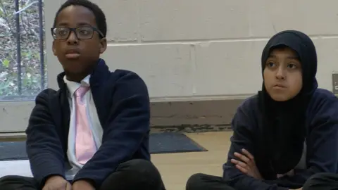 The image shows two school pupils in dark blue uniform.  On the left is an 11 year old boy, on the right a 10 year old girl.  They are sat cross legged on the floor watching the Life of a Chickpea schools session which is teaching them about equality, diversity and inclusion.