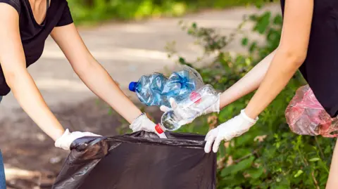 Two women are holding a black binbag between them. They both have white gloves on. The women on the right is putting two plastic bottles in the bag.