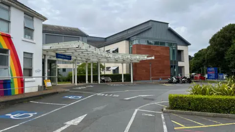 BBC Guernsey's Princess Elizabeth Hospital - A white building with a ranbow painted on it. Next to it is a large criss crossed awning, above a sign which says Le Vauqueidor Entrance. 