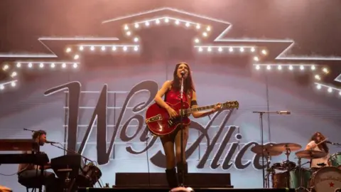 Reuters Wolf Alice performs at Open'er Festival in Gdynia, Poland July 5, 2025. Lead singer and guitarist Ellie Rowsell stands centre stage and plays a red guitar. The name of the band is displayed in large letters behind her.