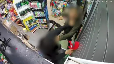 A CCTV image shows a shop assistant being assaulted inside a convenience store in Liverpool. A man whose face is blurred can be seen throwing a punch over the counter at the person working behind it.