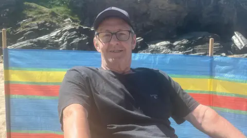 A man with glasses wearing a dark t-shirt and baseball cap sitting in front of a multi-coloured wind-break on the beach with rocks in the background