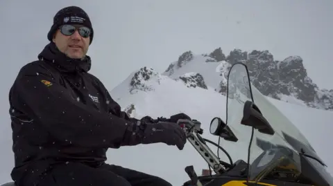 Supplied Prof John Woodward in black jacket and black sunglasses and black beanie. He is sitting on a snowmobile with snowy mountains in the background.