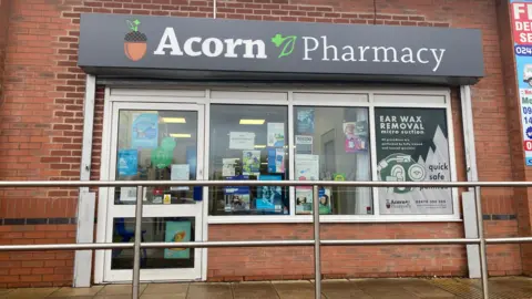 The outside of a pharmacy, called Acorn Pharmacy. There is a picture of an acorn on the signage, with medical posters in the window 