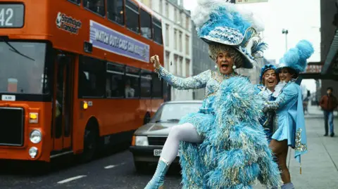 Stanley Baxter stands at a bus stop, dressed as a panto dame in a blue dress and large hat. He is sticking his leg out into the road comically, as a large red bus with a Strathclyde Transport sign drives by. Two other cast members stand behind him, laughing.