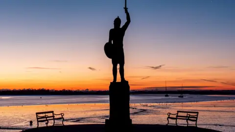 Mike Pountney A photo of Byrhtnoth statue in Maldon's Promenade Park, showing a warrior holding up a sword on his right arm and a shield in his left. It is in silhouette with a bench either side, a golden horizon and the Blackwater estuary in front, with two ships in the distance. 