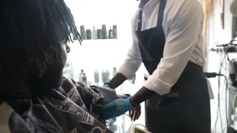 A staff member at Jul's barber shop in south London looks at a customer while testing his blood pressure with an out-of-shot machine that has a cuff