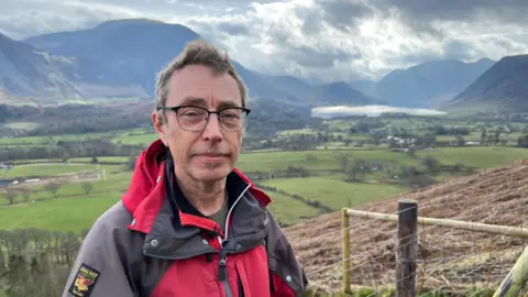 Darren Ward is standing on a hillside in Cumbria with Crummock Water and various fells visible in the background. He is wearing a red and grey coat and wearing glasses.