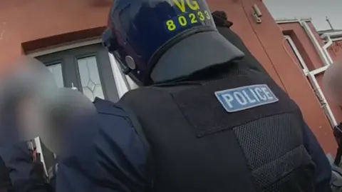 Body-cam footage shows a police officer from behind, dressed in protective gear, stood outside a front door