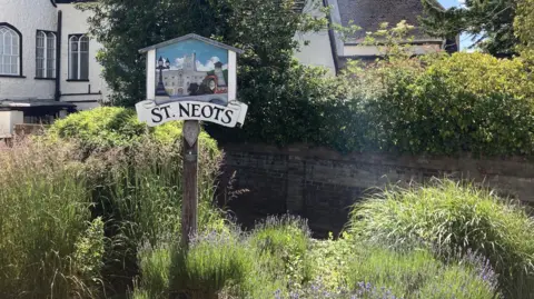 Emma Howgego/BBC A sign which is engraved with the words ST NEOTS in black font is on the left of the frame. It is positioned in between hedges and wildflowers. There is a large white house in the background.