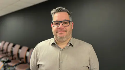 BBC Paul Bowring wears glasses and a gray shirt. He stands in front of a black wall.