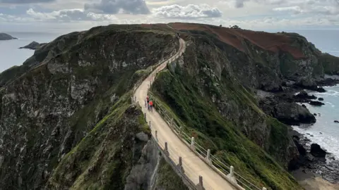 Sark's Coupeé - A winding bridge with rails on each side and green covered cliffs sloping towards sea on either side. On the bridge are two cyclists and at the end is a horse and cart.