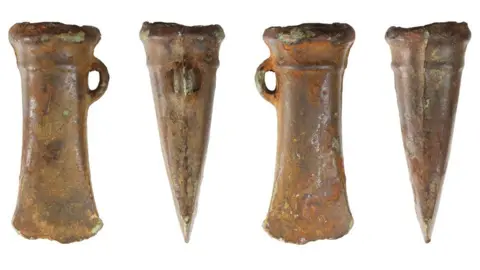 Andrew Williams/Norfolk County Council Four views of a bronze socketed axe head with double mouldings at the socket and a side loop. The cutting edge is slightly flared. The views all show the socket edge at the top and the cutting edge at the bottom. They show from left to right, the flat edge with the intact loop on the right; a side view with the loop on top; the flat edge with the intact loop on the left; a side view without the loop.  