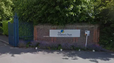 Google A red brick wall with a sign reading The Children's Trust adjacent to blue iron gates