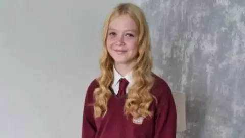 A young woman with long blonde hair, wearing a burgundy school uniform is seen smiling at the camera
