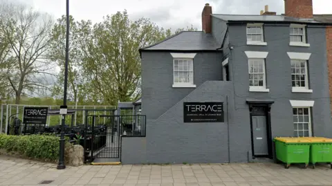 The outside of the Terrace bar. It is a grey building with an outdoor terrace garden, which has glass screens around it.