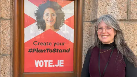 Emily Thomas standing in front of a poster which reads 'Create your #PlanToStand - Vote.je.' Emily is wearing glasses and has long hair.