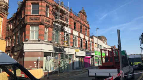 Scaffolding is on the left in front of two visible storeys of a building, not the ground floor. The blue sky is at the top of the image.