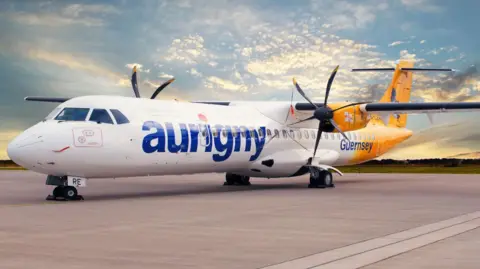 A yellow and white airplane with Aurigny logo on its side, setting off into the sky from the take off area at the airport. Blue skies and green grass visible.