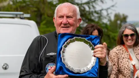 LS Photography A man with white hair stands holding a commemorative silver salver. He is wearing a black referees outfit.