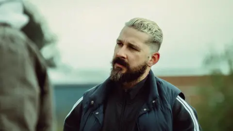 Shia Labeouf has bleached blonde hair slicked back and cut short on the sides. He wears an earring and has a dark brown long beard. He wears a black zip up top with two whitge stripes on the sleeves and a black gilet. 