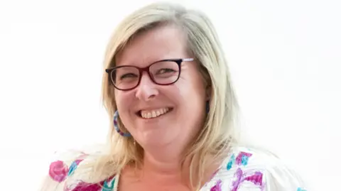 Anthony Cullen A head and shoulders image of Lucy Humphrey. She is wearing glasses, has long, light-coloured hair and is wearing a white blouse that features a bold, colourful pattern.