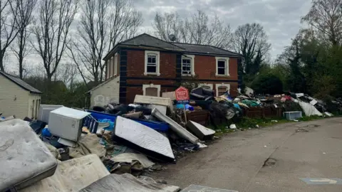 Aidan Kelly The old station, now with broken and boarded up windows. All along the road in front of it, and around the building, is flytipping - mattresses, white goods, blankets, and all kinds of rubbish. There is a red For Sale sign in front.