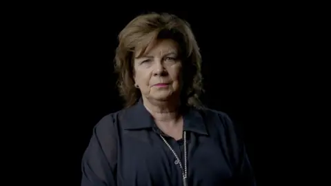 Elaine C Smith in black blouse and with pearls is standing against a blacj background, looking directly down the camera lens.