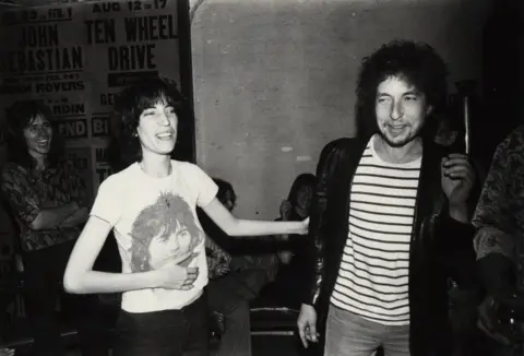 Chuck Pulin/Cache Agency Patti Smith on the left, wearing a white T-shirt with a photo of a man smiling on it. She is holding out her arm to Bob Dylan, on the right. He is wearing a white striped shirt and leather jacket.
