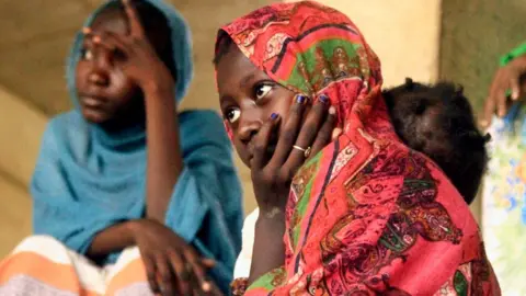 Two young women - one wearing a red headscarf and the other in a blue headscarf, sit at a shelter where they were evacuated by the Sudanese army in Omdurman - May 2025.