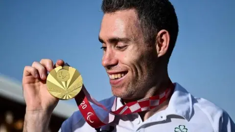 Getty Images Athlete Jason Smyth holds a gold medal. he is wearing a white track suit and is smiling broadly.