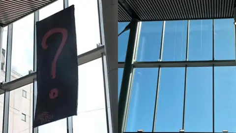 To the left is an image of a black banner with a large pink question mark on it hanging from a ceiling. To the right is an image of the same space without the question mark. 